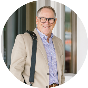 A photo of Ed Chaffin in brown tux and a light purple button down carrying a bag on his left arm while smiling for a photo
